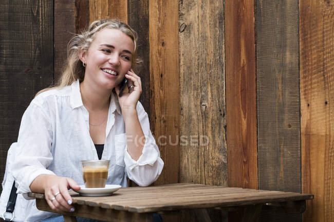 Jovem loira sentada sozinha em um café, usando telefone celular, trabalhando remotamente. — Fotografia de Stock