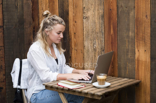 Young blond woman wearing face mask sitting alone at a cafe table with a laptop computer, working remotely. — Stock Photo