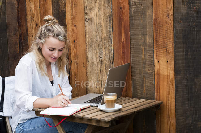 Young blond woman sitting alone at a cafe table with a laptop computer, writing in note book, working remotely. — Stock Photo