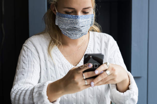 Молода блондинка в масці для обличчя сидить наодинці в кафе, використовуючи мобільний телефон, працює дистанційно . — стокове фото