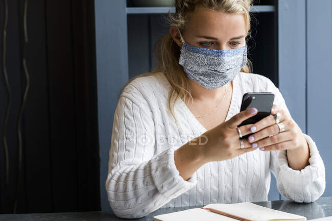 Giovane donna bionda che indossa maschera facciale seduta da sola in un caffè, utilizzando il telefono cellulare, lavorando a distanza. — Foto stock