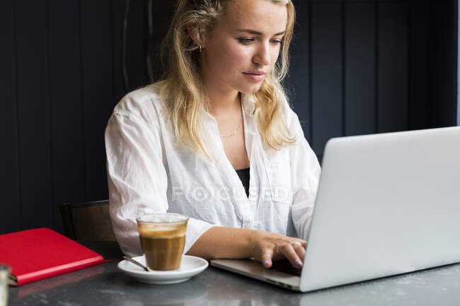 Mujer rubia joven sentada sola en una mesa de café con una computadora portátil, trabajando remotamente. - foto de stock