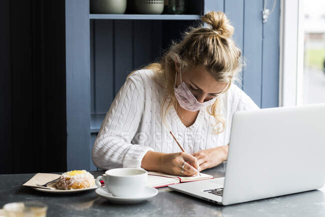 Mujer rubia joven con máscara facial sentada sola en una mesa de café con una computadora portátil, escribiendo en un cuaderno de notas, trabajando remotamente. - foto de stock