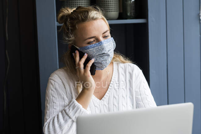 Junge blonde Frau mit Gesichtsmaske sitzt allein an einem Cafétisch mit einem Laptop-Computer, benutzt Mobiltelefon, arbeitet aus der Ferne. — Stockfoto