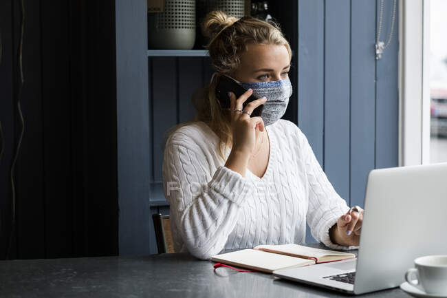 Mujer rubia joven con máscara facial sentada sola en una mesa de café con una computadora portátil, usando un teléfono móvil, trabajando remotamente. - foto de stock