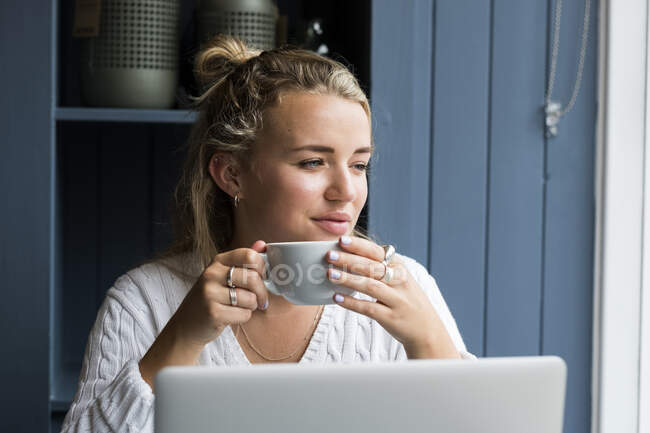 Young blond woman sitting alone at a cafe table with a laptop, working remotely. — Stock Photo