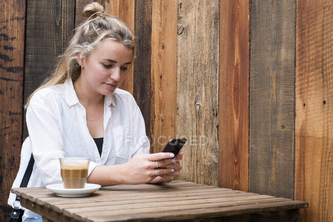 Junge blonde Frau sitzt allein an einem Café-Tisch im Freien und checkt ihr Handy. — Stockfoto