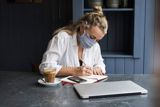 Жінка носить маску для обличчя наодинці за столом кафе з ноутбуком і блокнотом, що працює віддалено . — стокове фото