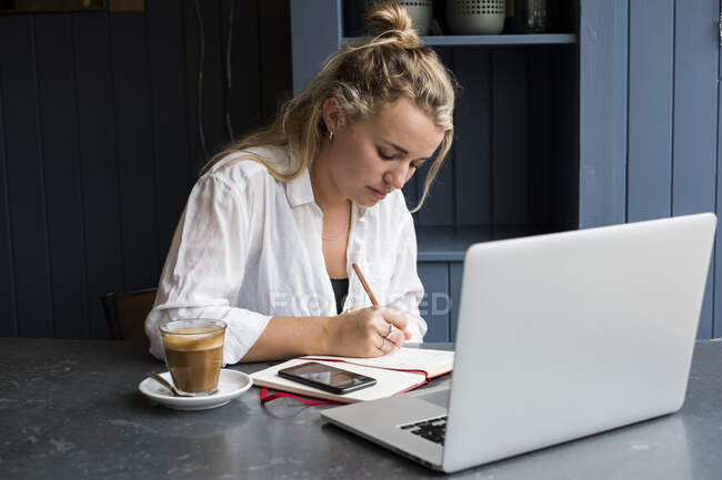 Frau sitzt allein mit Laptop am Cafétisch, schreibt Notizbuch, arbeitet aus der Ferne. — Stockfoto