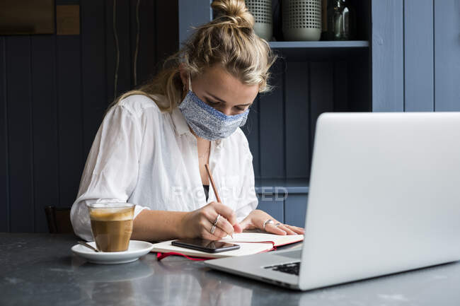 Femme portant un masque facial assise seule à une table de café avec un ordinateur portable, écrivant dans un carnet de notes, travaillant à distance. — Photo de stock