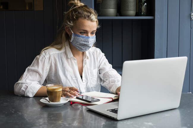 Женщина в маске для лица сидит одна за столиком кафе с ноутбуком, пишет в записной книжке, работает дистанционно. — стоковое фото