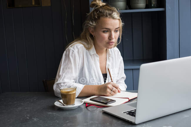Жінка сидить наодинці за столом кафе з ноутбуком, пише в записнику, працює дистанційно . — стокове фото