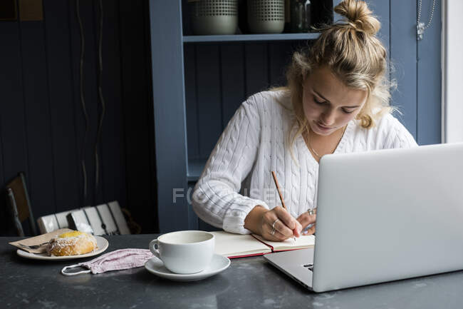 Mujer sentada sola en una mesa de café con un portátil, escribiendo en un cuaderno, trabajando remotamente. - foto de stock