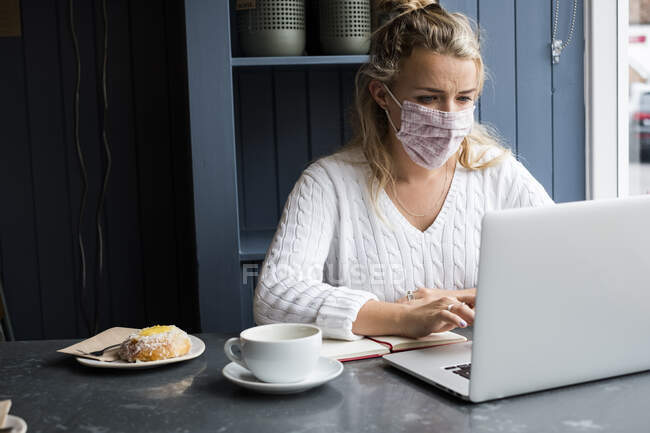 Mujer con máscara facial sentada sola en una mesa de café con un portátil, escribiendo en un cuaderno, trabajando remotamente. - foto de stock