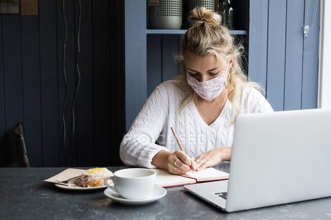 Frau mit Gesichtsmaske sitzt allein an einem Cafétisch mit Laptop, schreibt in Notizbuch, arbeitet aus der Ferne. — Stockfoto