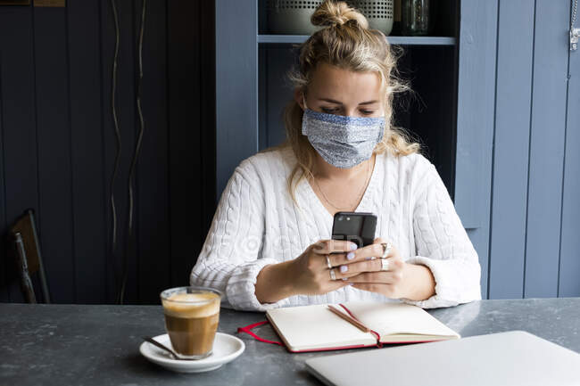 Жінка в масці на обличчі сидить на самоті за столом у кафе з ноутбуком, користується мобільним телефоном і віддалено працює.. — стокове фото