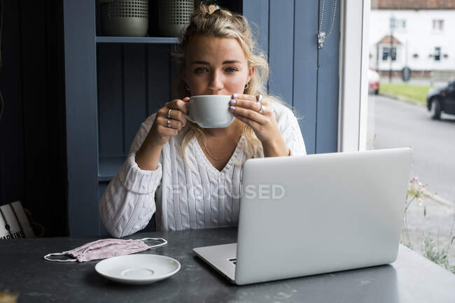 Jeune femme blonde assise seule à une table de café avec un ordinateur portable et une tasse de café — Photo de stock