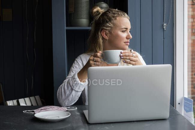 Mujer rubia joven sentada sola en una mesa de café con una computadora portátil, y una taza de café - foto de stock
