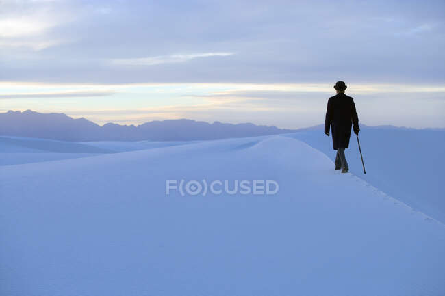 Vista posteriore dell'uomo in cappotto e cappello a bombetta passeggiando attraverso un paesaggio duna bianca con un bastone. — Foto stock
