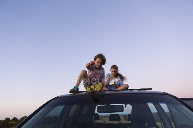 Adolescente et son jeune frère assis sur la voiture SUV au coucher du soleil. — Photo de stock