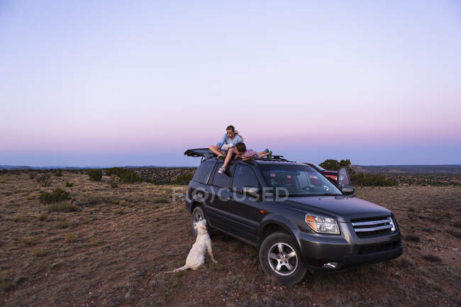 Teenage girl and her younger brother sitting atop SUV car at sundown. — Stock Photo