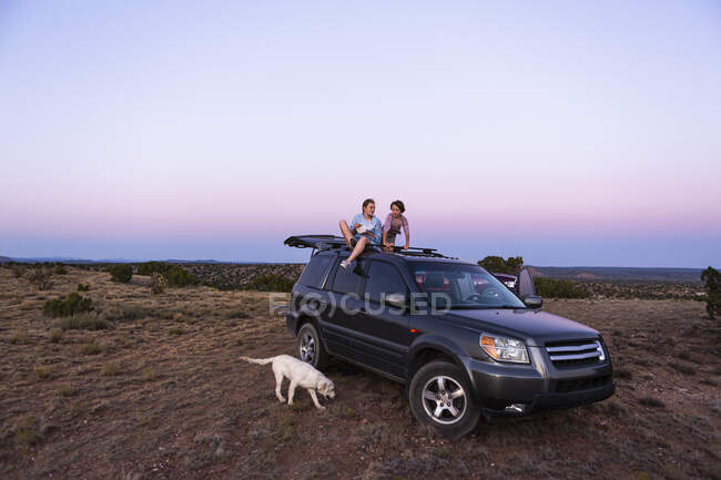 Teenage girl and her younger brother sitting atop SUV car at sundown. — Stock Photo
