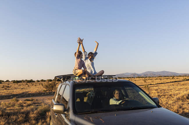 Teenage girl and her younger brother on top of SUV car driving on desert road — Stock Photo