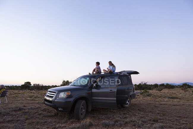 Adolescente et son jeune frère assis au sommet de leur SUV au coucher du soleil. — Photo de stock