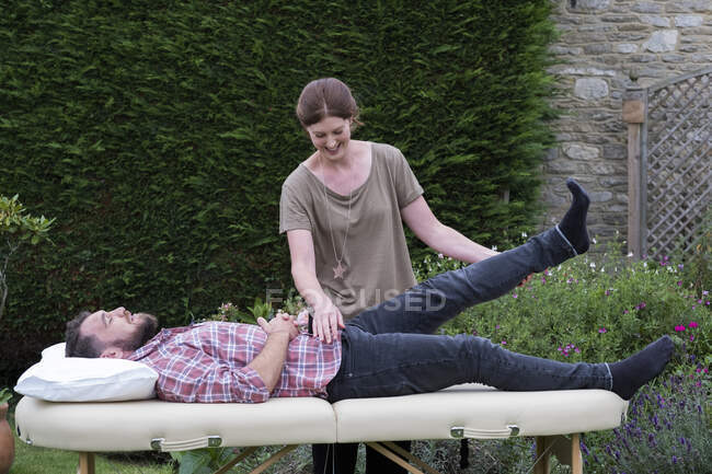 Homme sur un canapé et thérapeute levant la jambe, dans une séance de thérapie de jardin — Photo de stock