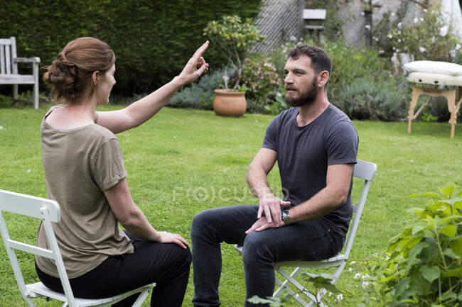 Terapeuta y cliente sentados en un jardín, mujer con la mano levantada y dos dedos extendidos. - foto de stock