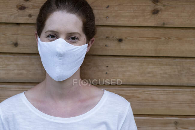Portrait of woman wearing white face mask, leaning against wooden wall, looking at camera. — Stock Photo