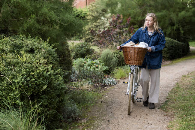 Mujer rubia joven empujando bicicleta con cesta a lo largo del sendero. - foto de stock