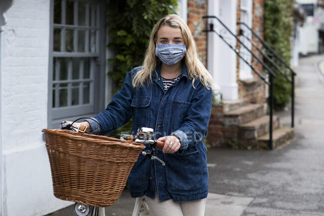Young blond woman wearing face mask on bicycle with basket, looking at camera. — Stock Photo