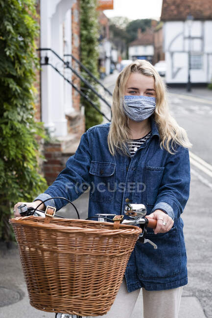 Mujer rubia joven con máscara facial en bicicleta con cesta, mirando a la cámara. - foto de stock