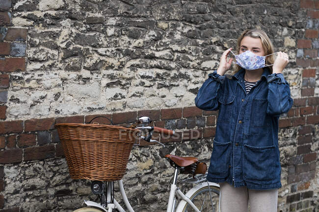 Junge blonde Frau steht mit Korb neben Fahrrad und setzt Gesichtsmaske auf. — Stockfoto