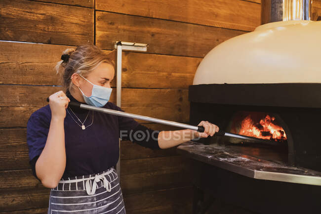 Blond waitress wearing blue striped apron and face mask standing at a pizza oven in a restaurant. — Stock Photo