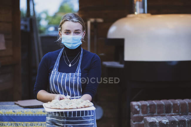 Mujer camarera en delantal y máscara facial sosteniendo pizza fresca en un tablero - foto de stock