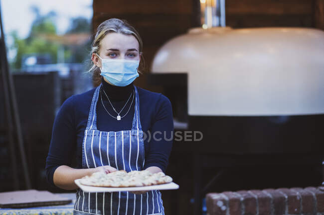 Kellnerin in Schürze und Gesichtsmaske hält frische Pizza auf einem Brett — Stockfoto