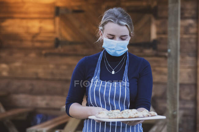 Mujer camarera en delantal y máscara facial sosteniendo plato de pizza. - foto de stock