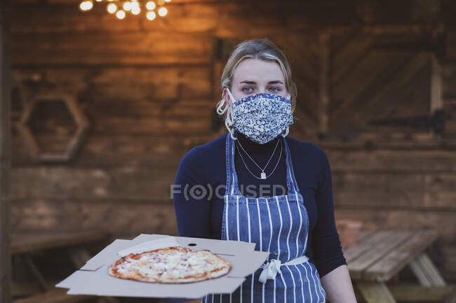 Cameriera donna in grembiule e maschera viso che tiene piatto di pizza. — Foto stock
