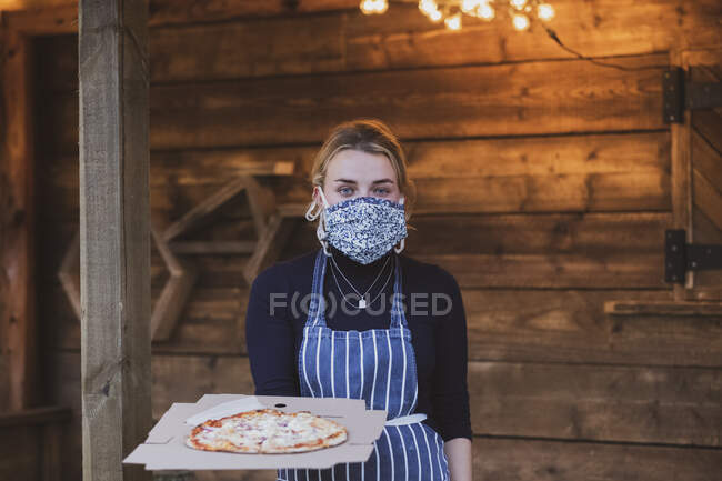 Kellnerin in Schürze und Mundschutz hält Teller mit Pizza. — Stockfoto