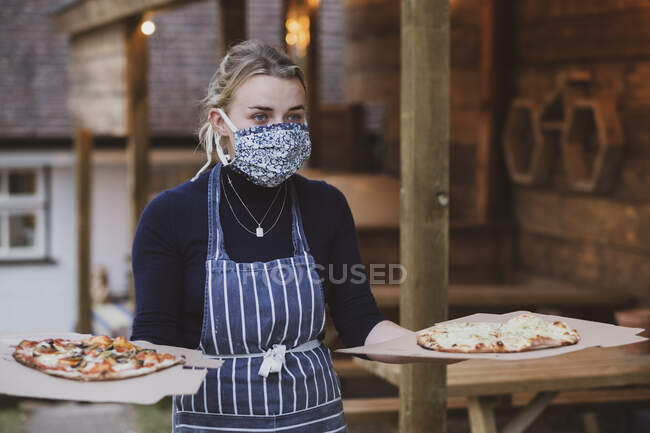 Mujer camarera en delantal y máscara facial sosteniendo platos de pizza. - foto de stock