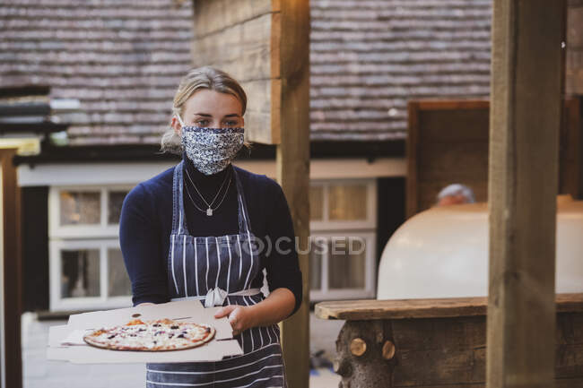 Официантка в фартуке и маске для лица с тарелкой пиццы. — стоковое фото