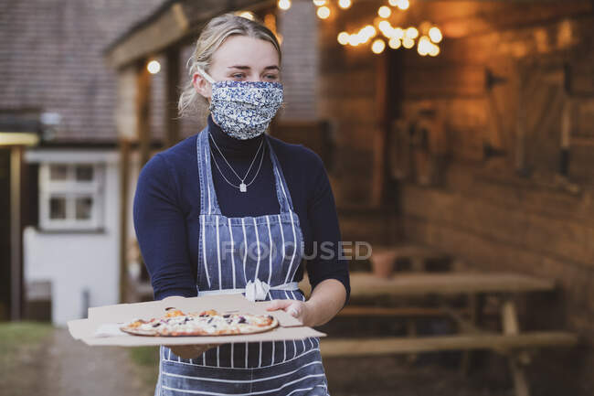 Mulher garçonete em avental e máscara facial segurando prato de pizza. — Fotografia de Stock