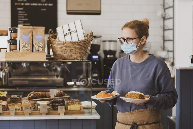 Camarera rubia con máscara facial trabajando en un café, llevando platos de comida. - foto de stock