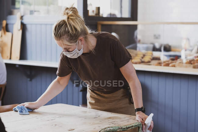 Blonde Kellnerin mit Mundschutz arbeitet in einem Café, wischt Tisch. — Stockfoto