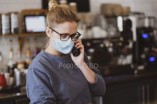 Serveuse blonde portant un masque facial travaillant dans un café, au téléphone. — Photo de stock