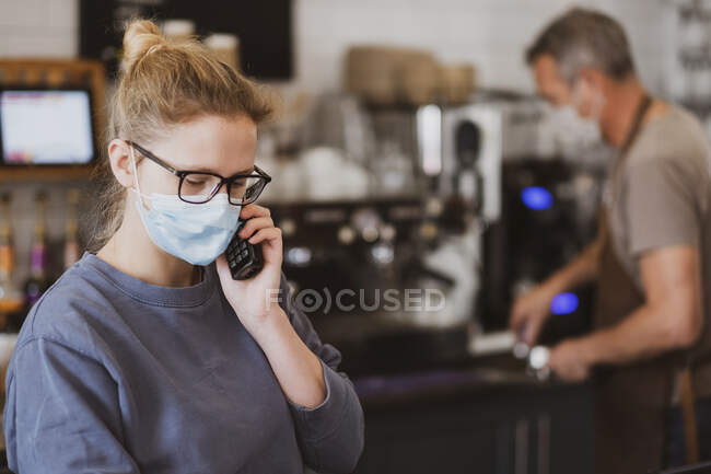 Camarera rubia con máscara facial trabajando en un café, por teléfono. - foto de stock