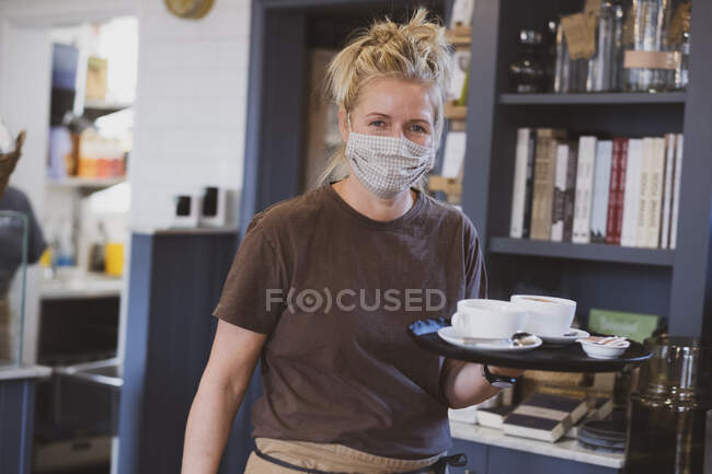 Garçonete loira usando máscara facial trabalhando em um café, transportando bandeja com xícaras de café. — Fotografia de Stock