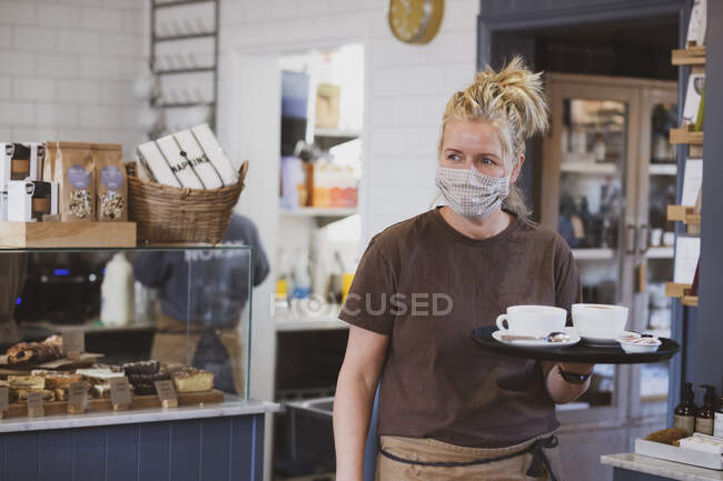 Camarera rubia con máscara facial trabajando en un café, llevando bandeja con tazas de café. - foto de stock